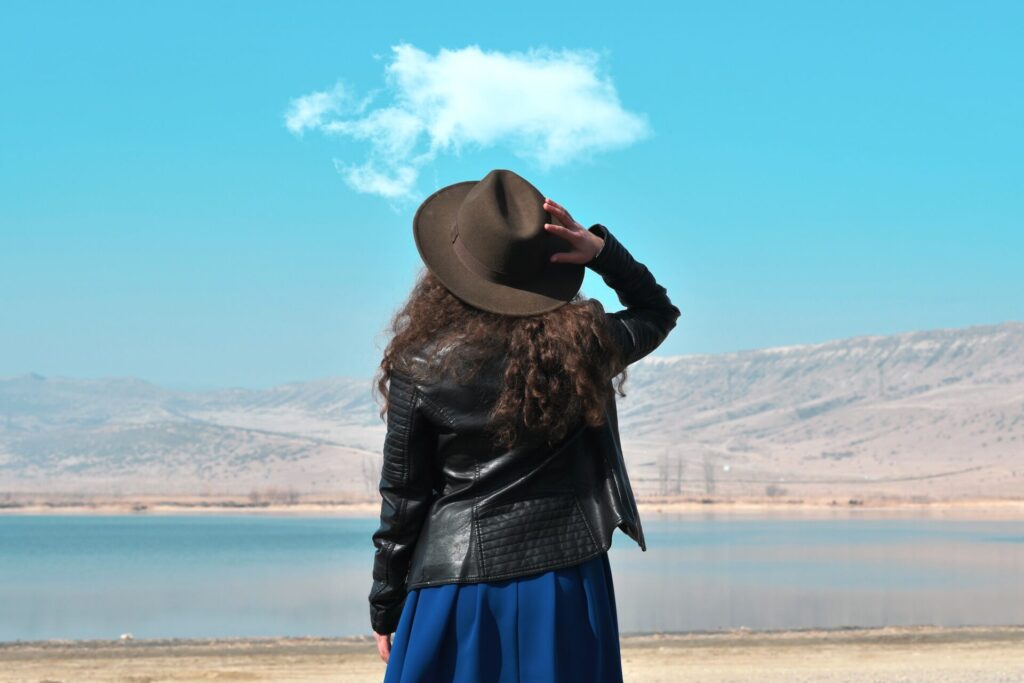 woman standing with her back turned, in blue skirt and black hat, looking toward a desert lake on the horizon. the sky is blue with one wispy white cloud.